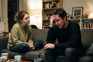 Homme interrompt dispute sur canapé, larmes aux yeux, partenaire observe avec empathie attentive.