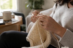 Mains d'une femme en train de tricoter avec de la laine crème et des aiguilles en bois, dans un salon lumineux et chaleureux