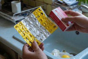 Mains tenant boîtes d’amoxicilline et Doliprane près d’un évier de cuisine.